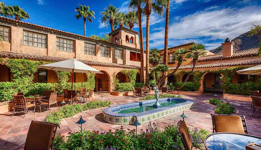 The gorgeous courtyard at the Royal Palms is seen on a bright day, umbrella-covered table and a mosaic tile lined fountain inviting visitors to relax