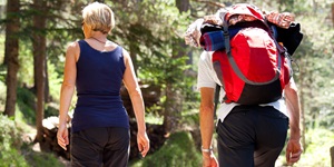 A woman in a blue tank top and leisure shorts and a man wearing a large red hiking backpack with sleeping gear hike in a woody area 