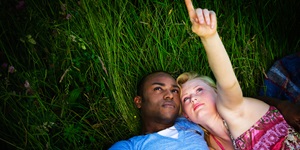 A man in a blue shirt and a woman in a tank top lie in the grass at night, the woman pointing up at the sky