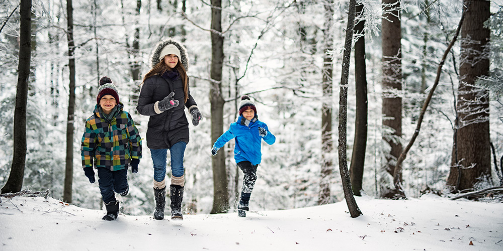 Mom and two boys run happily through a wintery forest, fresh white snow covering the area as the three of them look happily bundled in their winter clothing