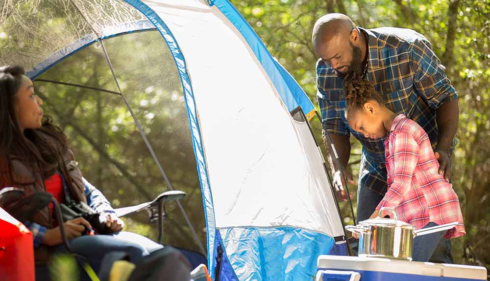 Woman sits watching a man and little girl setting up a tent