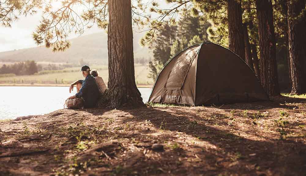 A pair of campers sit by the edge of a lake by their tent in the woods