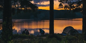 Three people hanging around a camp fire with a tent by the lake at sunset