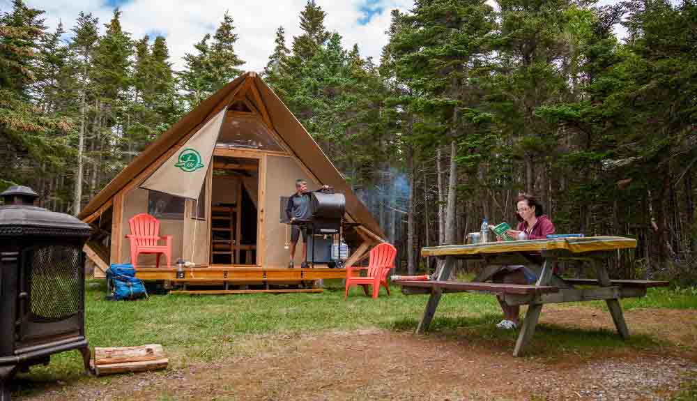 Glamping site with a man barbecuing as a woman reads at a picnic table