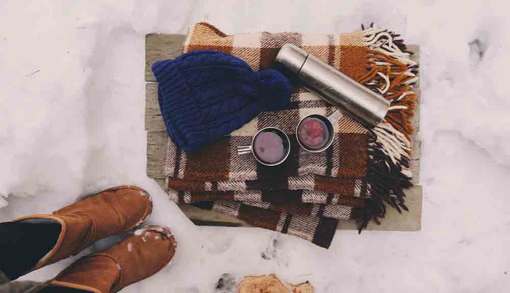 A picnic blanket is shown on the snow with a thermos and mugs