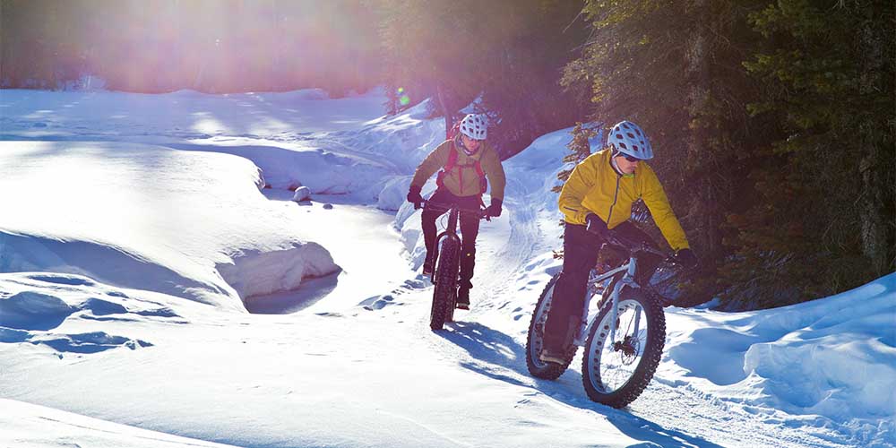 Two people are shown riding fat tire bikes through the snow with trees in the background