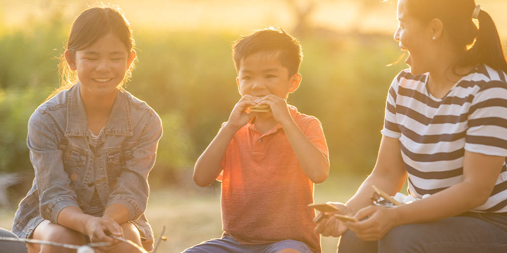 A woman and two kids eat s'mores at sunset
