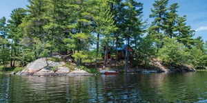 A cottage is shown on a rocky shore with trees all around it and water in the foreground