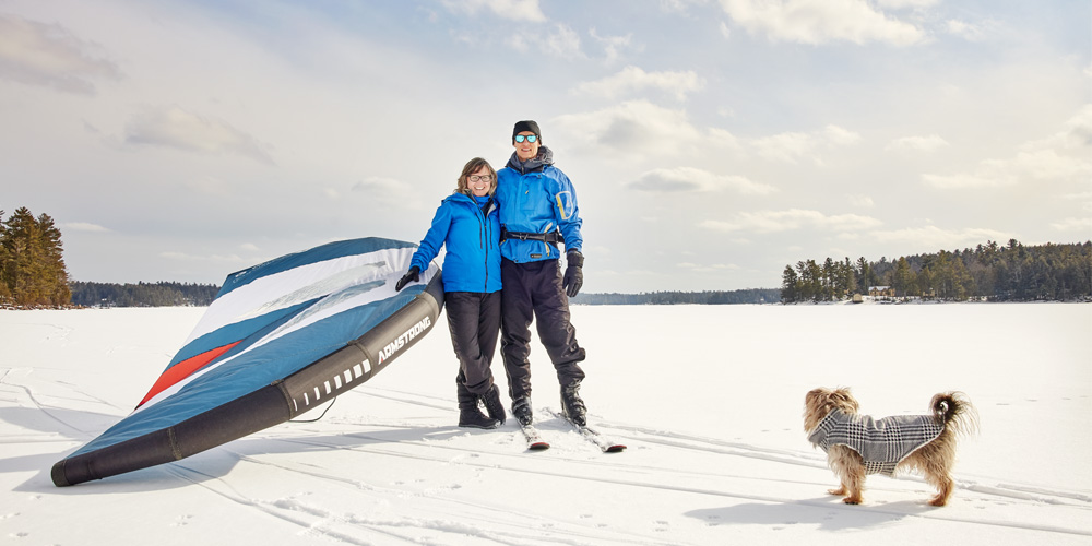 Two people in black snow pants and bright blue winter jackets stand in an empty snowy field. The person on the left is shorter than the one on the right and they are holding a large kite. With them is a small brown dog.