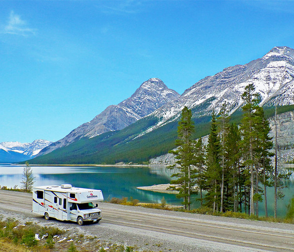 A white RV drives on a road beside mountains and a clear, blue lake.