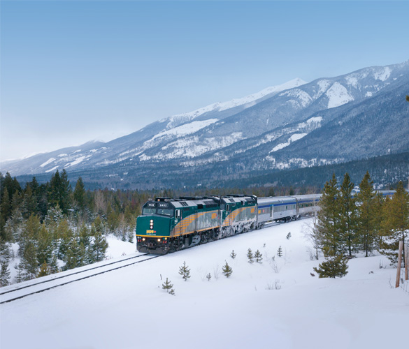A branded VIA Rail train going through a snowy forest with mountains in the background.