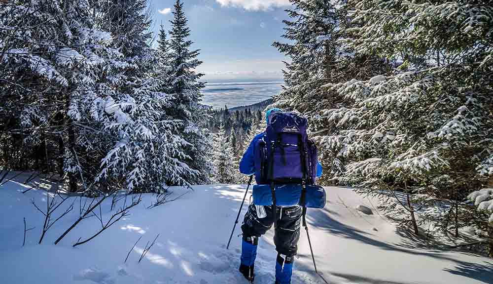 Person in winter coat and snow pants with a loaded backpack as they snowshoe up a hill 