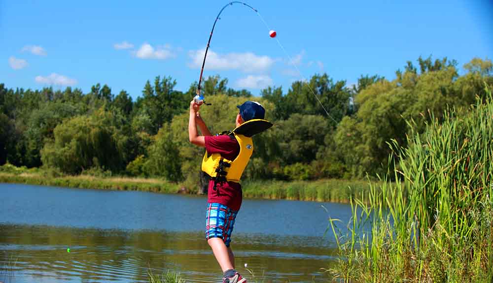 Little boy wearing yellow lifejacket with a fishing rod in the air by a pond in the summer