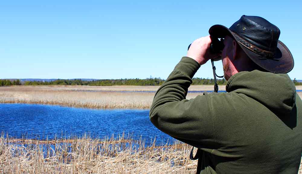 Man wearing green sweatshirt and a leather cowboy hat looking through binoculars