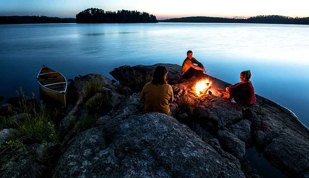 Three people sitting on a rock by the water of Thunder Bay around a small fire