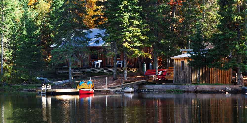 A camping lodge is seen on a calm lake with a canoe tied to the dock, wood boat house near the shoreline and large wooden lodge seen partially hidden among the trees behind