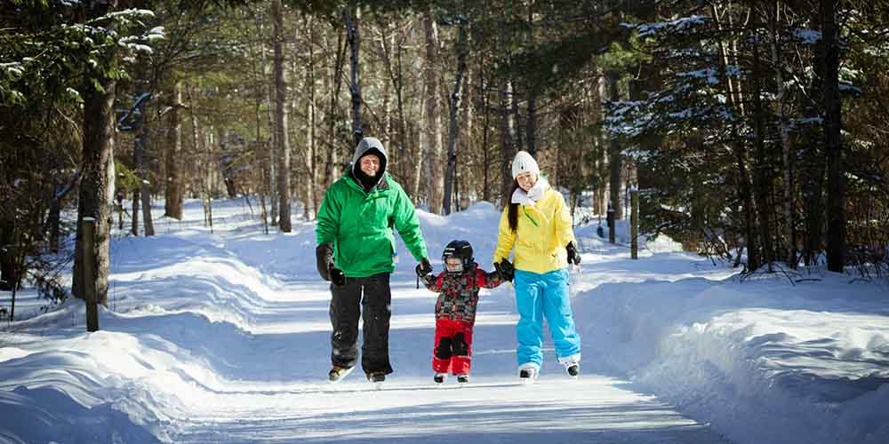 A man, woman and child skate hand-in-hand down a snowy path in Arrowhead Provincial Park while wearing bright cozy winter jacks, hats, gloves and snow pants
