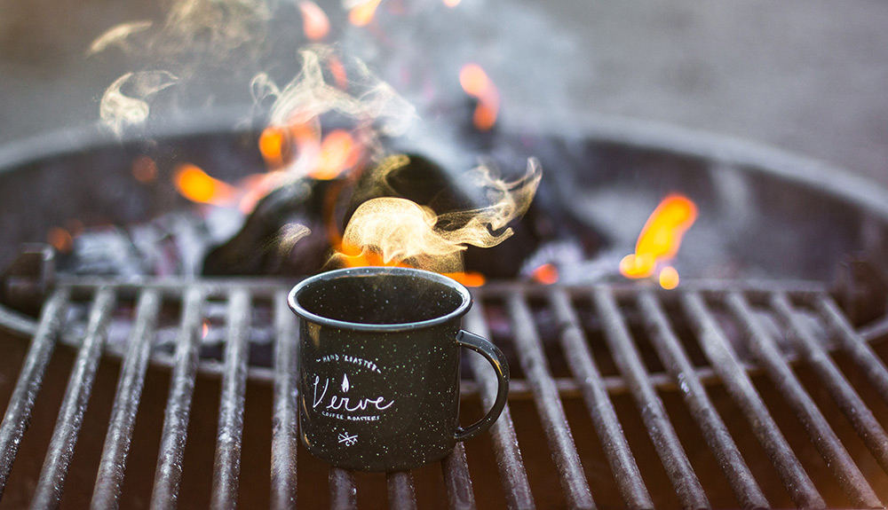 A camping mug sits atop a grill in front of a blazing campfire