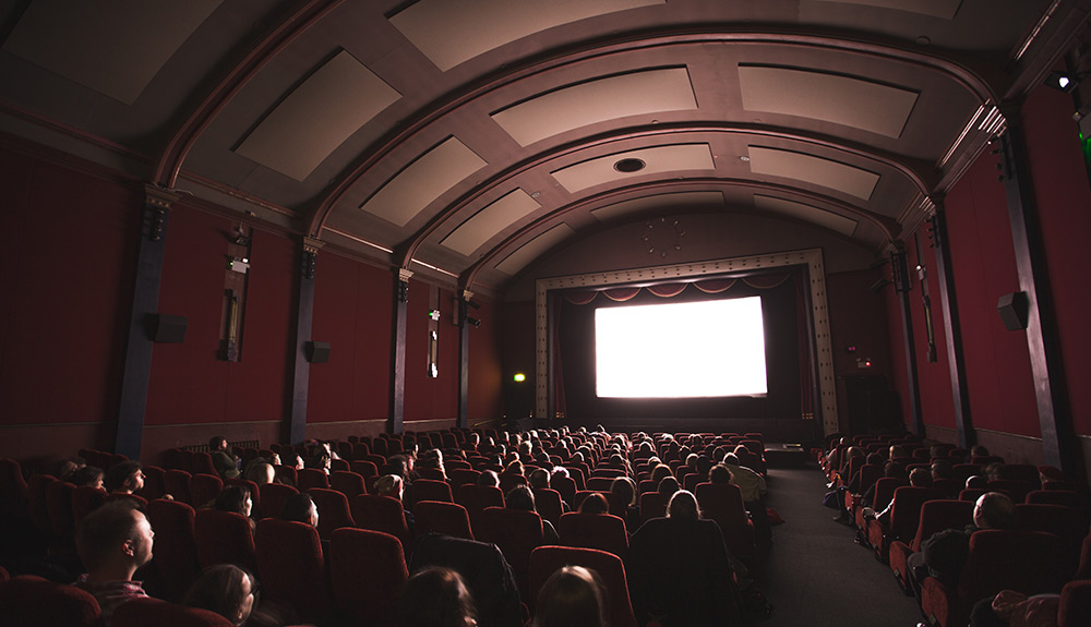 A dimly lit movie theatre is shown full of people sitting in their seats