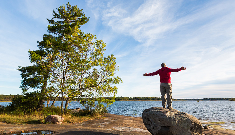 A man stands on a large rock, arms outstretched as he gazes out at Lake Muskoka