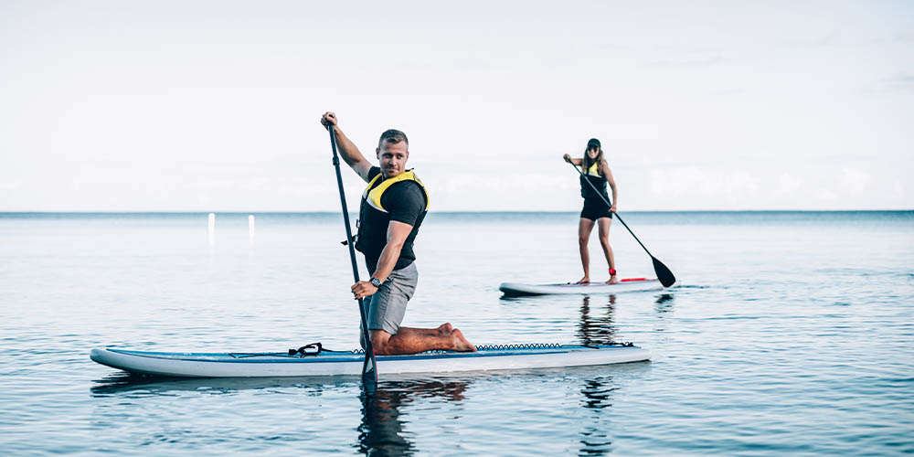 There are two people on the water on paddleboards. There is a man wearing grey shorts, a black T-shirt and a black and yellow life jacket. He is kneeling on a white and blue standup paddleboard, holding a black paddle that is partly submerged in the water. Behind him, several feet away, is a woman with long blonde hair, wearing a black baseball cap and black shorts, and the same black and yellow life jacket. She is standing up on her white paddleboard and is smiling at the camera with her sunglasses on.