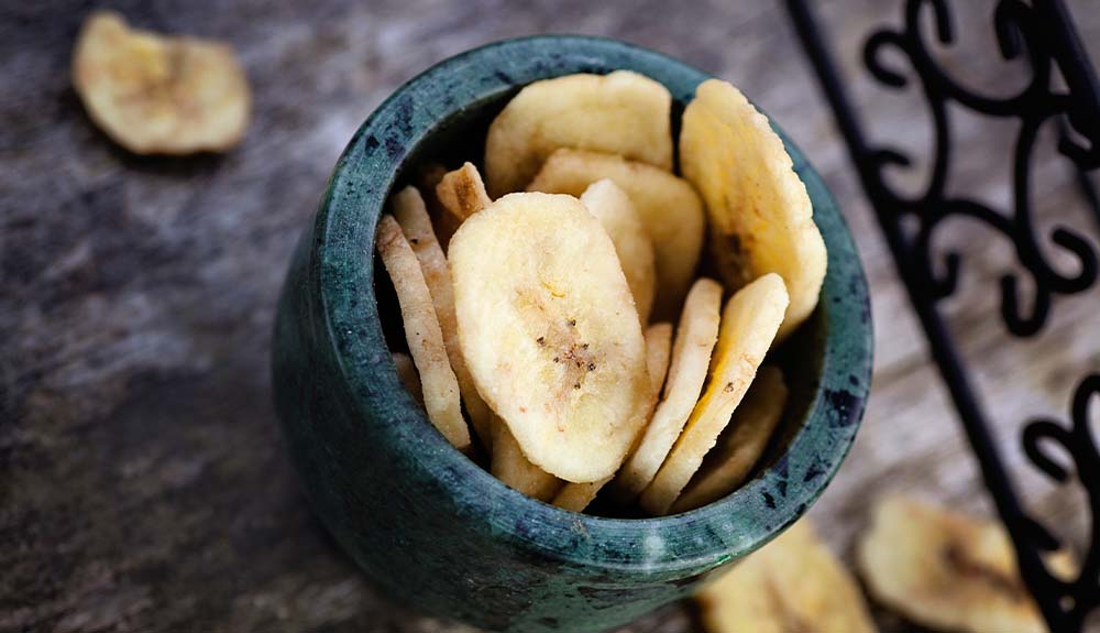 A small green and black bowl filled with banana chips that are spilling over onto the table.