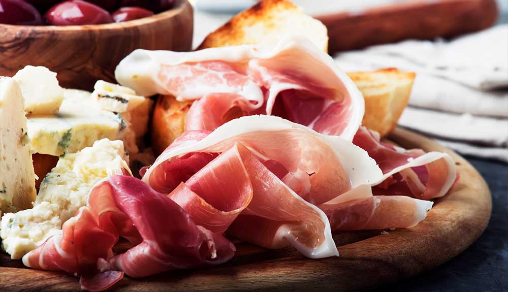 A wooden tray piled with some chunks of grilled bread, cured meats like prosciutto and cheese.