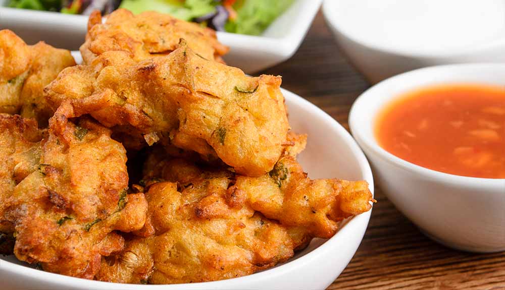 A white bowl filled with crispy vegetable pakora and an orange dipping sauce next to it in a separate white bowl.