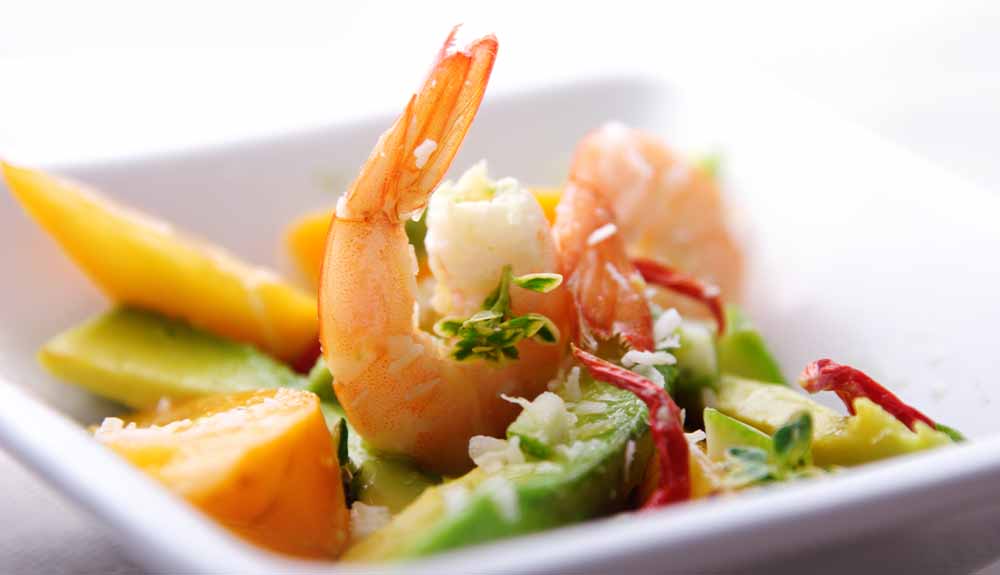 A square dish with a single shrimp placed in the middle with its tail up surrounded by some sliced fruits and vegetables.