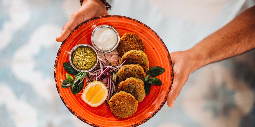 Two hands holding out a bright orange plate with five crispy falafels on the side, with a lemon cut in half on the side, with two round bowls with dip, one that is white and one that is yellow with green in it.