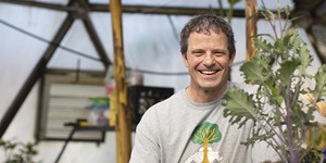 Science teacher Steve Schultz wears a grey t-shirt and stands next to a plant in a greenhouse with windows showing a blue sky behind him.
