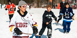 Children wearing hockey jerseys and holding hockey sticks while playing the sport together on an outdoor rink. There is snow covering the ground around the ice and there are trees in the background.