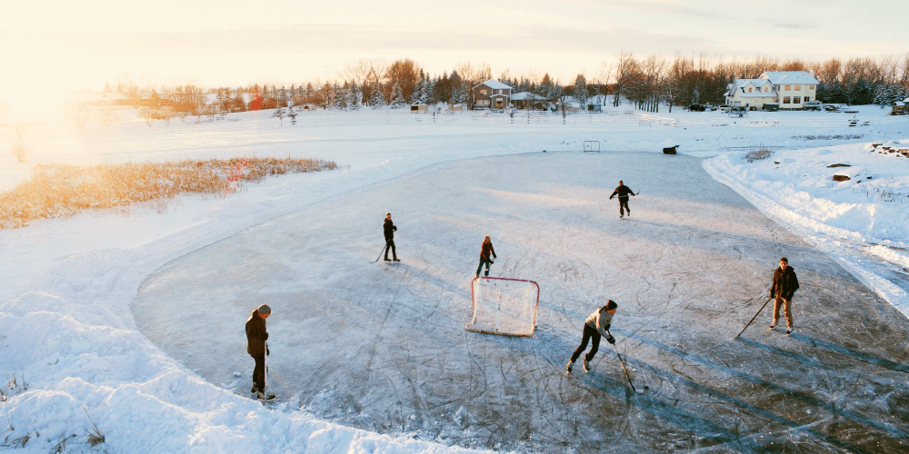 An outdoor ice rink surrounded by a field covered in snow. There are people on the ice skating around, with a hockey net placed at one end. The sun is setting in the background.