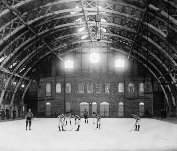 An indoor ice rink with high ceilings, in black and white. There are a few people skating on the ice and there are 3 bright lights above them.