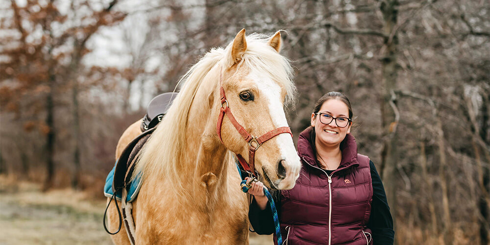 WETRA Owner Paige Shepley wears a black long sleeve shirt with a purple puffer best and stands next to a light brown horse with a white mane.
