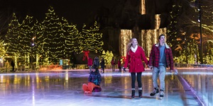 A couple ice skating at night. There are evergreen trees in the background decorated with lights. 