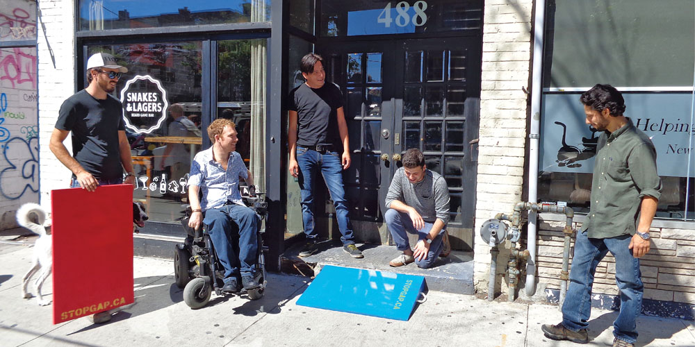 A group pf people, one of which is in a wheelchair, stand in front of a shop and look down at a bright blue wheelchair ramp that connects the sidewalk to the store's entrance.