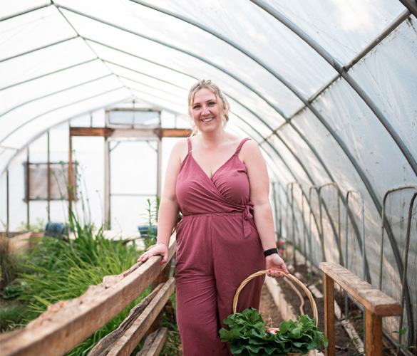 A woman in a mauve jumpsuit holds a brown basket full of produce. She is standing inside a greenhouse with a clear covering and rows of produce growing.