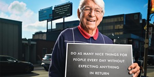 An older man wearing a blue sweater over a red collared short, with grey hair and round metal glasses, holds a black sign that says "Do as many nice things for people every day without expecting anything in return."