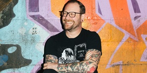 A man wearing a black tshirt and black glasses stands in front of a vibrantly coloured graffiti wall with his arms crossed.