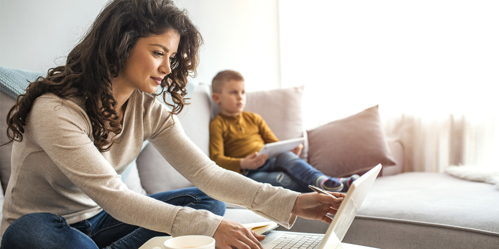 A woman with long, brown hair and a grey long sleeve shirt sits on a beige couch with a laptop open in front of her. On the other side of the couch sits a small boy wearing a yellow long sleeve shirt.
