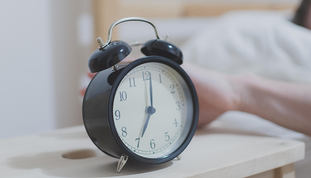 A black alarm clock sits on a bedside table indicating the time is seven AM