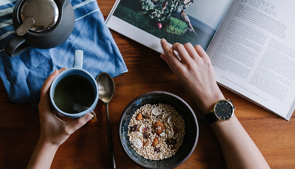 A hand holds a mug of coffee, the other hand flips the page of a magazine, a bowl of muesli laid out on the table