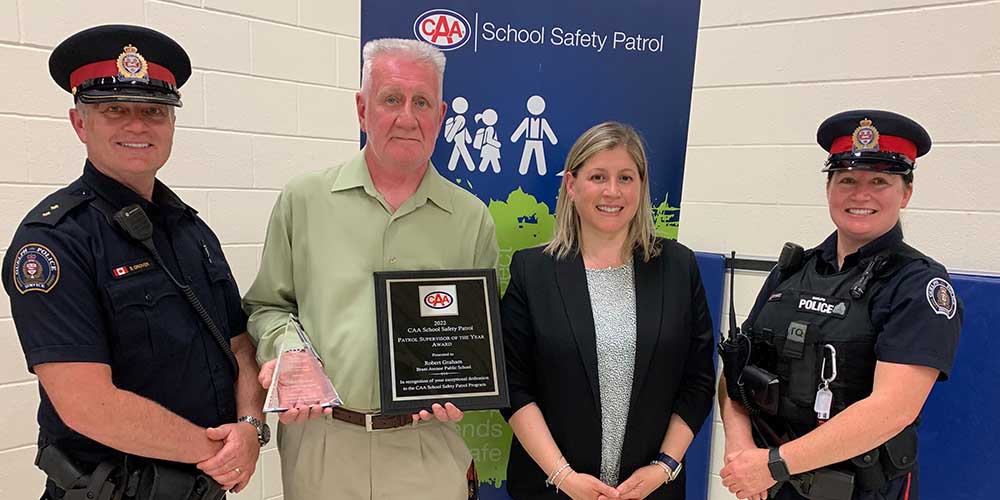 From the left: a Guelph police officer wearing his black uniform and hat with red and gold police emblem; Robert Graham, winner of the CAA School Safety Patrol Supervisor Award, wearing a green polo and beige khaki pants, holding a triangular glass trophy and a plaque; a woman wearing  a grey and white speckled shirt and a black blazer; a Guelph police officer wearing her uniform and hat with red and gold police emblem as well as a chest protector with a radio, walkie-talkie, flashlight and keys. Behind them is a white brick wall and a dark blue and white CAA School Safety Patrol banner. 