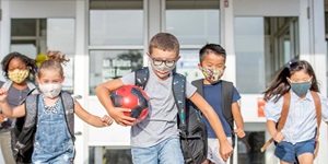 Five school-aged kids leaving the front entrance of a building, each wearing backpacks and a mask. The boy in the centre is wearing glasses and is carrying a red and black soccer ball. 