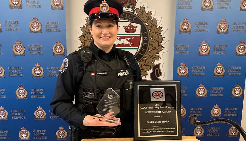 A police officer in full uniform with a black cap with a red ribbon around the middle, a black long-sleeved, shirt and a black vest is standing at a podium. On her vest is a small Canadian flag embroidered on it. She is holding a glass award and a black and gold plaque. Each have the CAA logo on it. Behind her is a wall with a dark blue background with a gold and white logo and the words Guelph Police Service repeated.
