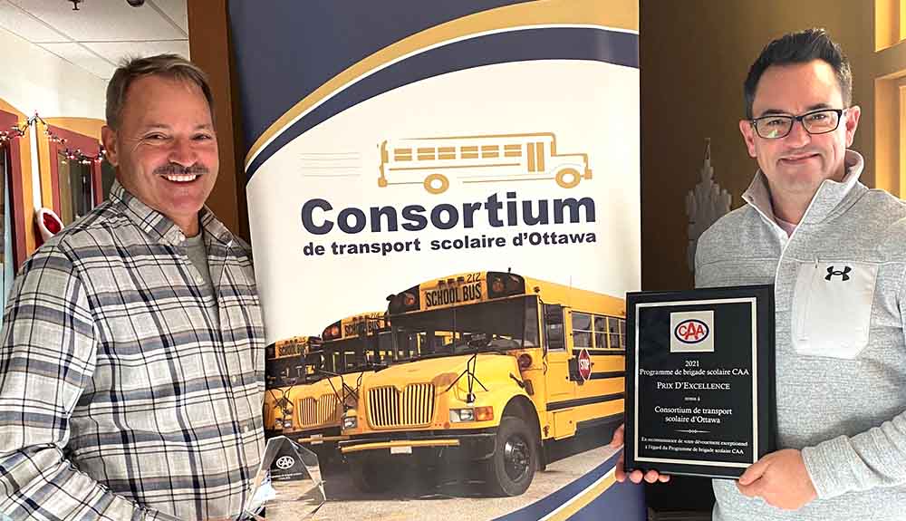 Two men standing in front of a sign that says Consortium de transport scolaire d'Ottawa. The man on the left is wearing jeans and a grey plaid shirt. He is holding a small, clear glass triangle with the CAA logo at the top. On the other side of the sign is a man wearing a light grey zip up shirt with jeans. He is holding a black plaque with the CAA logo on it. 