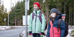 Two young kids are standing outside. There is a taller girl standing on the left. She is wearing a mint green coat with a red ear warmer and black gloves. She is wearing a backpack with purple straps and a gray mask. Next to her is another person wearing a navy and pink colour block winter coat with a navy knit hat. That person is wearing a black backpack with a print on it. 