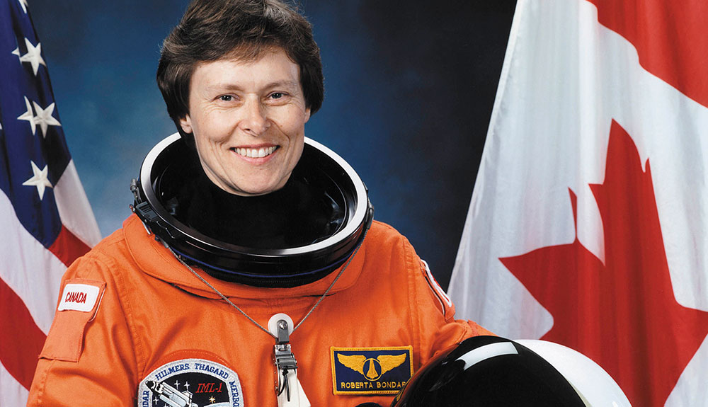Dr Roberta Bondar wearing an orange space suit while posing in front of American and Canadian flags in a 1992 photograph