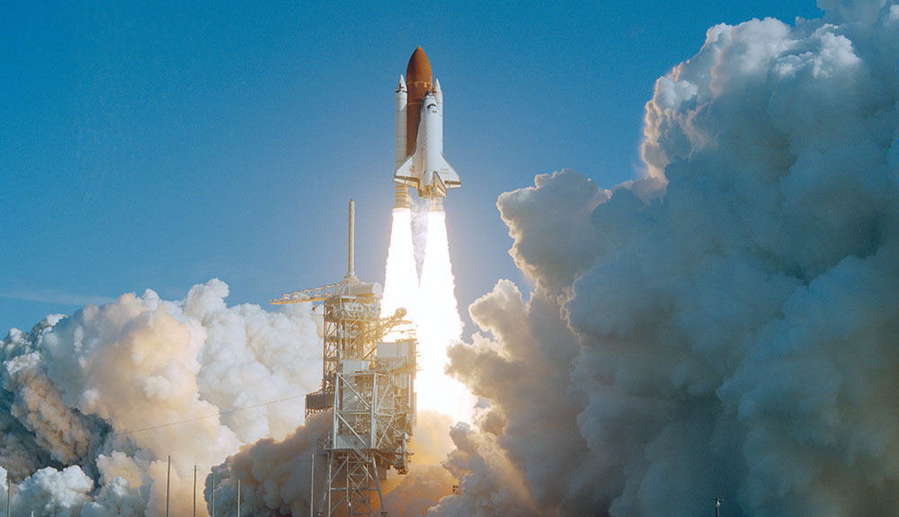 Discovery makes its ascent into space, its engines flaring behind it and large plumes of smoke covering the area